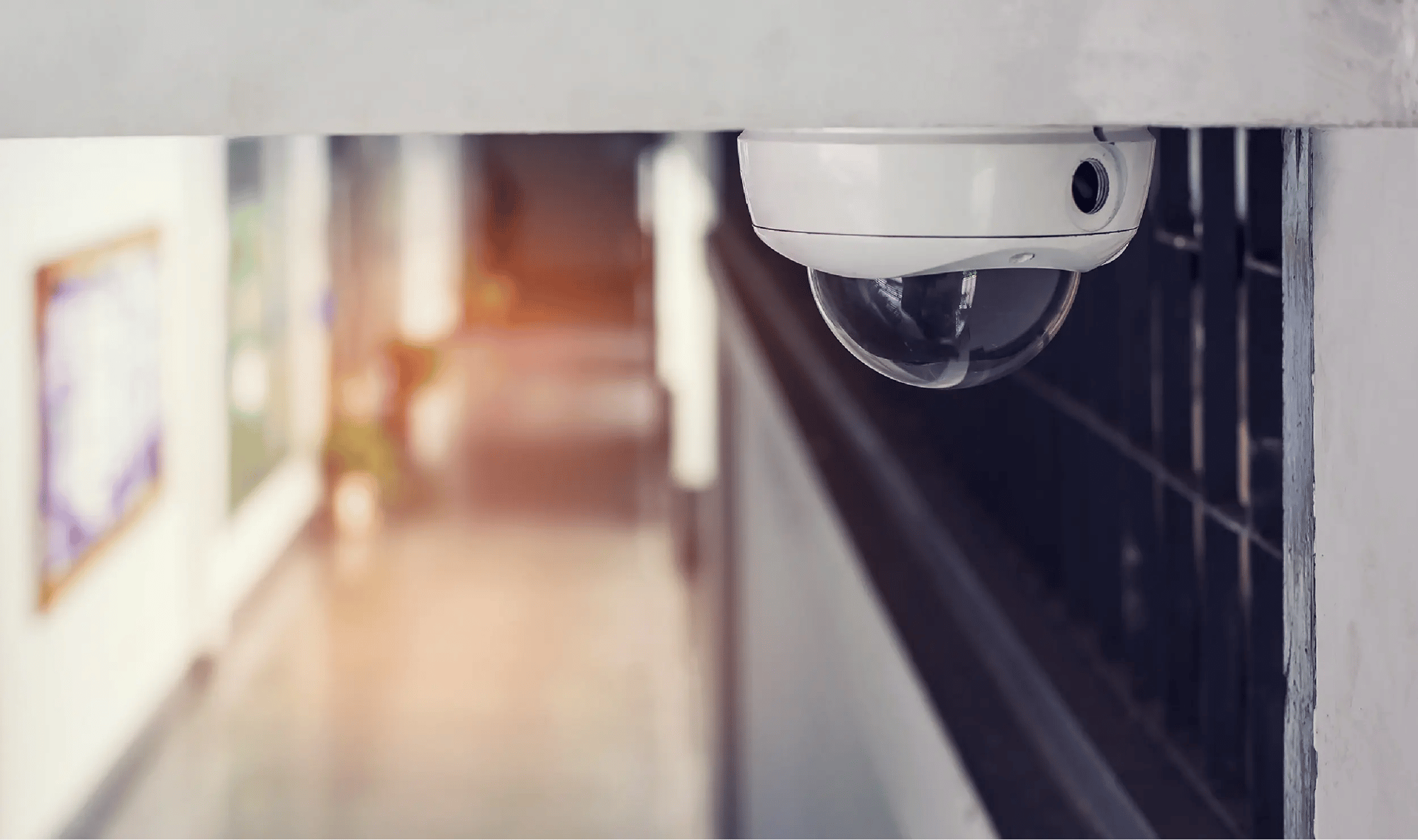 Close-up of a dome security camera mounted on the ceiling inside a commercial building, overlooking a blurred hallway to represent indoor video surveillance and physical security monitoring. Close-up of a dome security camera mounted on the ceiling inside a commercial building, overlooking a blurred hallway to represent indoor video surveillance and physical security monitoring.