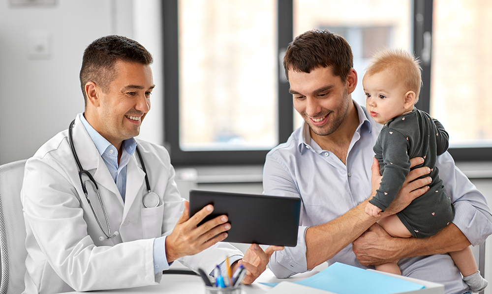 A smiling doctor in a white lab coat with a stethoscope shows a tablet to a father holding a baby during a pediatric clinic appointment.