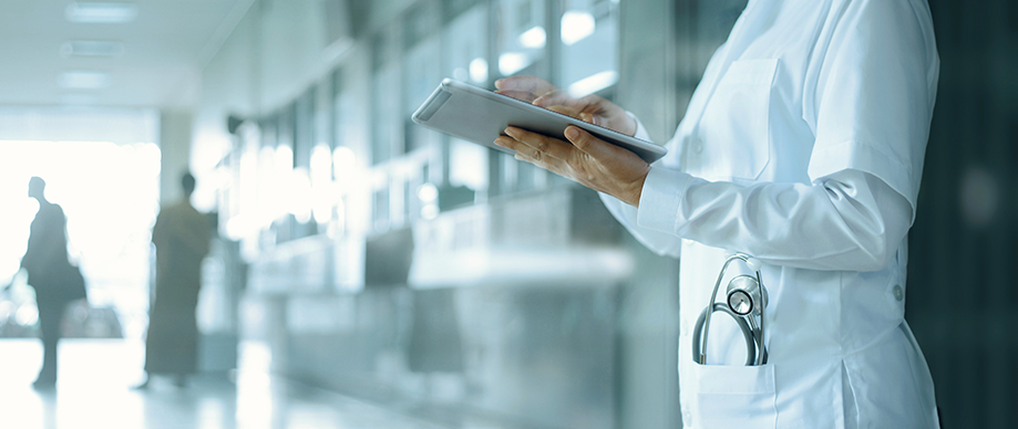 Healthcare professional in a white lab coat holding a tablet in a modern hospital corridor, with a stethoscope visible in their coat pocket.