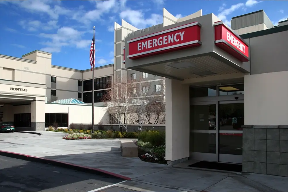 Exterior of a hospital emergency entrance with red emergency signs, automatic sliding doors, an American flag, and a clear blue sky. Exterior of a hospital emergency entrance with red emergency signs, automatic sliding doors, an American flag, and a clear blue sky.