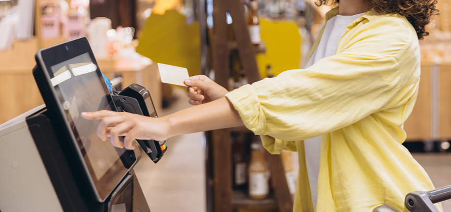 Shopper tapping a touchscreen self-checkout kiosk while holding a payment card at a convenience store.