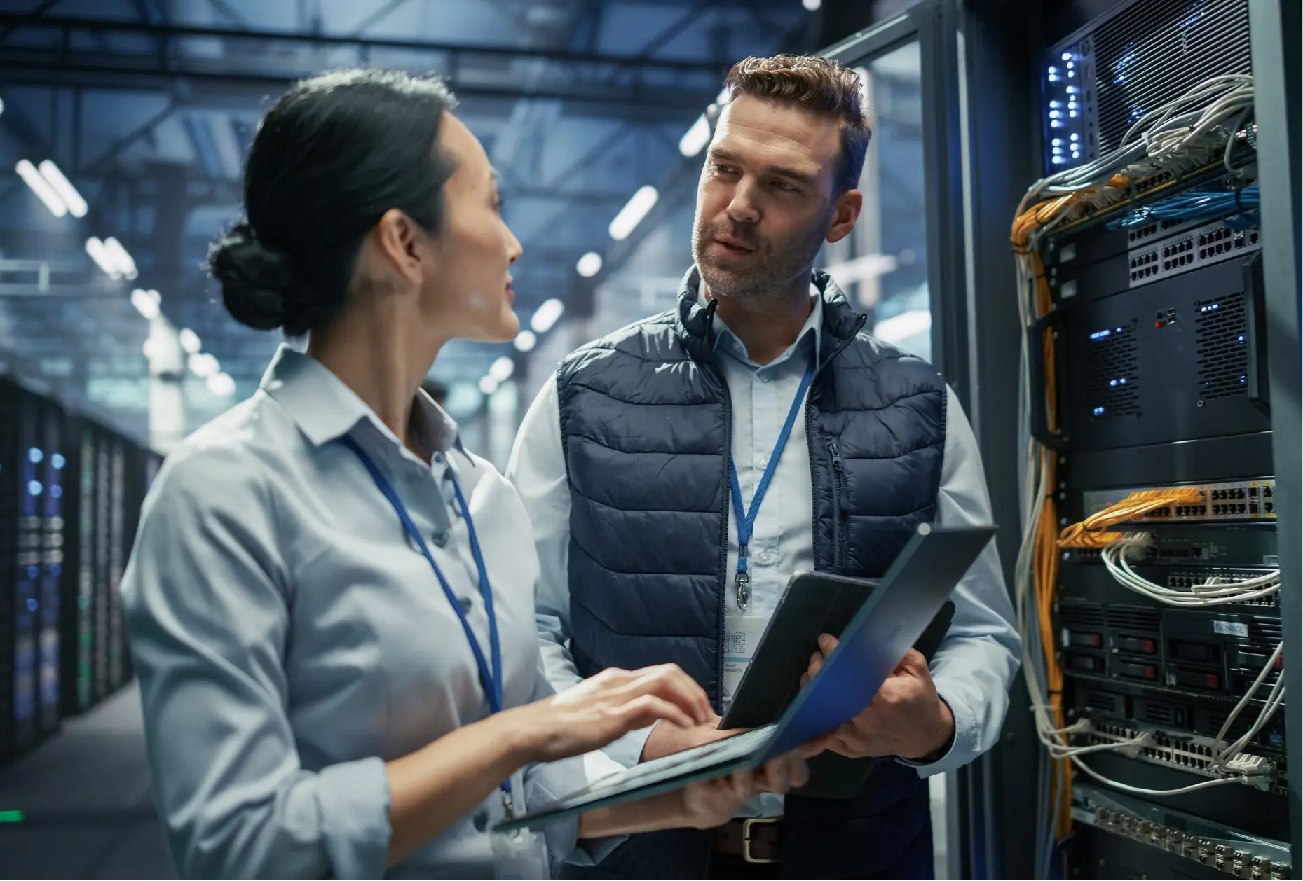Two IT professionals reviewing network infrastructure on a laptop and tablet in a data center server room, discussing technology sourcing strategy.