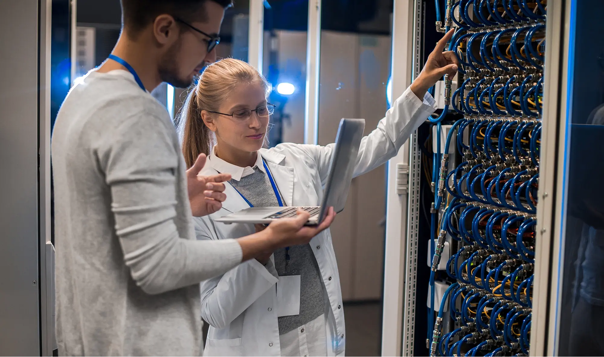 Two IT technicians holding a laptop and inspecting cable connections inside a modern server room rack. Two IT technicians holding a laptop and inspecting cable connections inside a modern server room rack.