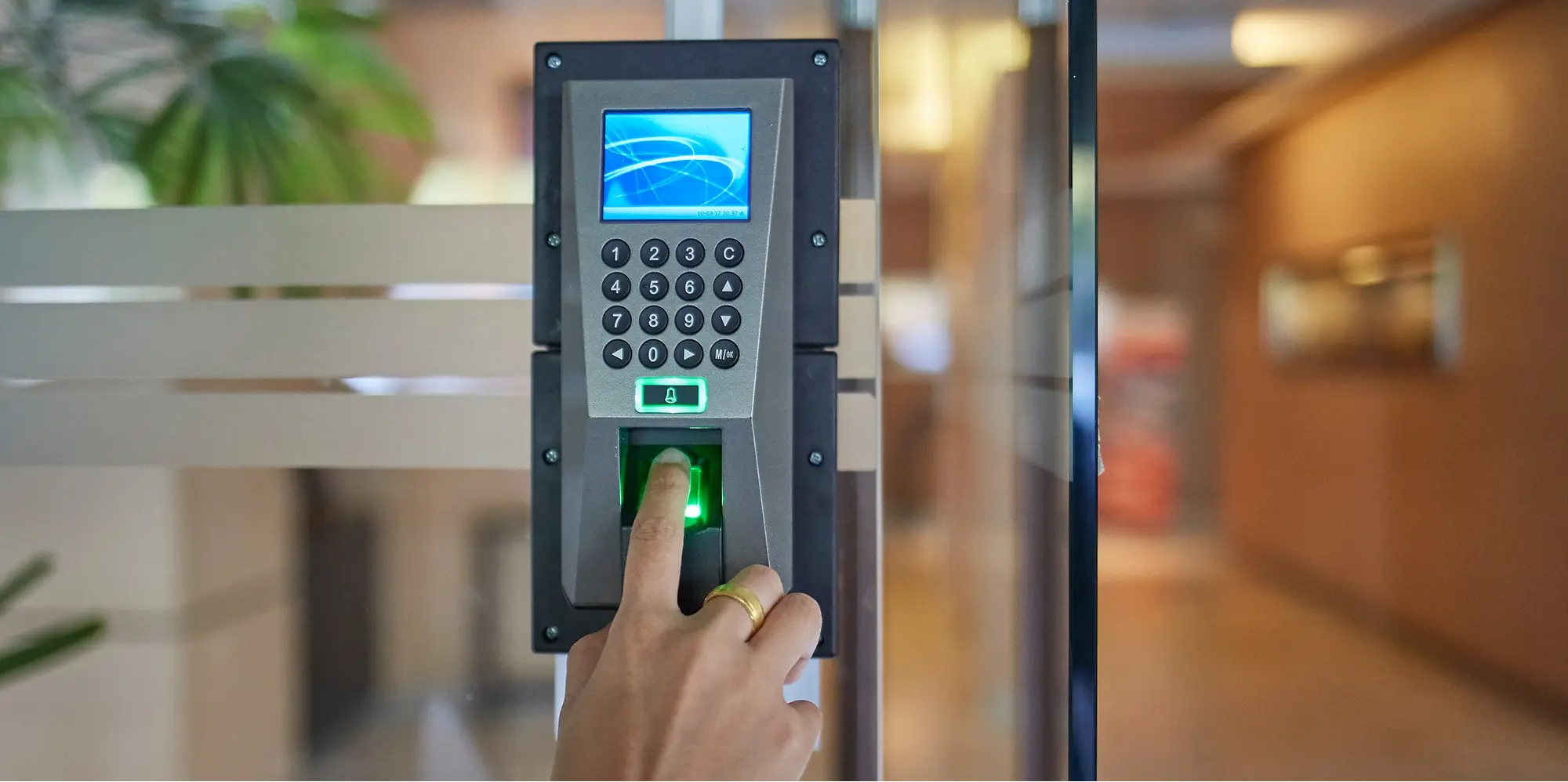 Person using a fingerprint access control system to unlock a secure glass office entrance, with a keypad and digital display mounted on the door.